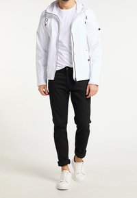 Mężczyzna ubrany w białą kurtkę na zamek, białą koszulkę, czarne podwinięte spodnie i białe trampki, stojący na neutralnym tle.