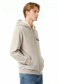 Hoodie zippé gris avec une doublure en sherpa crème, présentant des cordons de serrage, deux poches avant et un texte imprimé sur la poitrine. Coupe décontractée.