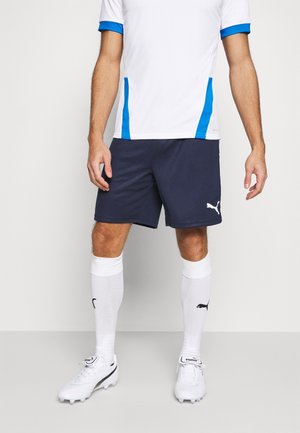 Männlicher Sportler trägt weißes und blaues Sporttrikot, marineblaue Shorts, weiße knielange Socken und weiße Puma Fußballschuhe und steht auf einem grauen Boden.