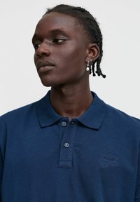 Polo majica temno modre barve z teksturirano tkanino, dvema gumboma na zapiranju in izvezenim logotipom na levi strani prsnega koša. Ima klasičen dizajn ovratnika.