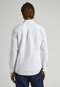 Camisa blanca de manga larga con rayas verticales en azul marino, puños abotonados, cuello estándar y dobladillo ligeramente curvado. Tejido texturizado.