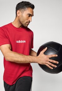 Punane puuvillane T-särk valge "adidas" logoga, mustade spordipükstega. Teema hoiab matimust meditsiinipalli.