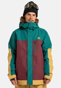 Chaqueta de esquí en tonos verde azulado, burdeos y mostaza con un alto cuello, cremallera frontal y bolsillo en el pecho. Presenta un diseño a bloques de color y una textura de tejido.