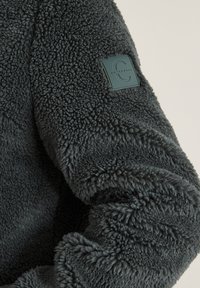 Chundelatá tmavě šedá fleecová bunda s texturovaným povrchem a obdélníkovou černou nášivkou na horní části rukávu, na které je vyobrazeno logo a název značky.