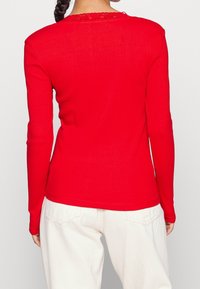 Punainen pitkähihainen ribbipusero, jossa on pyöreä pääntie ja koristeellinen pitsireuna selässä, yhdistettynä valkoisiin housuihin.