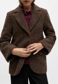 Brun uldblandingsfrakke med tekstureret overflade, med klippede lapels, enkelt knaplukning og brede ærmer, parret med en lilla skjorte under.