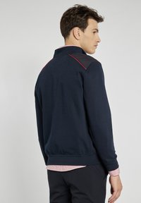 Granatowy sweter z kołnierzem stojącym, kontrastującymi czarnymi panelami na ramionach, czerwonymi detalami i prążkowanymi mankietami oraz dołem. Teksturowany materiał.