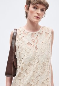 Δαντελένιο τοπ σε κρεμ απόχρωση με περίτεχνα στροβιλισμένα σχέδια και λουλουδάτα διακοσμητικά στοιχεία. Διαθέτει στρογγυλή λαιμόκοψη και σχέδιο χωρίς μανίκια, συνδυασμένο με καφέ τσάντα.