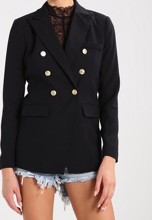 Mujer vestida con un blazer negro de doble botonadura con botones dorados sobre un top de encaje negro y shorts vaqueros claros desgastados.