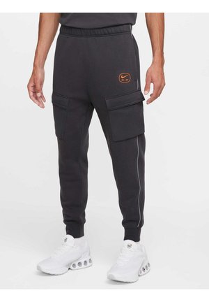 Mand iført sorte Nike Air cargo-sweatpants med elastiske manchetter og hvide træningssko på en ensfarvet baggrund.
