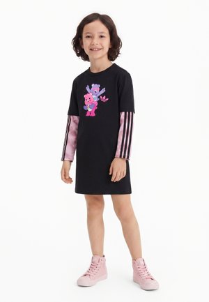 Enfant souriant, portant une robe noire avec des ours roses dessinés, des manches longues roses à rayures noires et des baskets montantes roses, debout sur fond blanc.