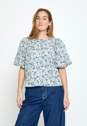 Femme debout portant un chemisier à manches courtes à imprimé floral avec des manches bouffantes et un jean bleu taille haute sur un fond blanc uni.