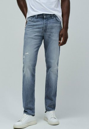 Hombre con vaqueros azules descoloridos con un ligero desgaste en el muslo izquierdo, combinados con zapatillas blancas y una camiseta blanca.