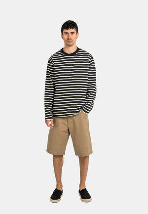 Mann steht, trägt ein schwarz-weiß gestreiftes Langarmshirt, beige knielange Shorts und schwarze Slipper vor weißem Hintergrund.