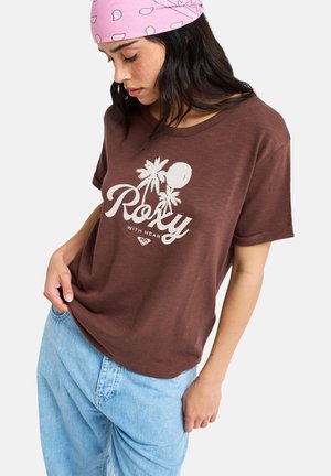 Jeune femme portant un t-shirt marron Roxy avec un motif de palmier, un jean bleu clair et un bandana rose sur la tête, regardant vers le bas.