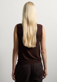 Top sans manches marron en tissu doux, avec un col rond et une coupe légèrement ajustée, associé à un pantalon foncé et des cheveux longs et raides.