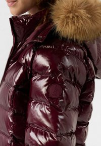 Giubbotto imbottito lucido bordeaux con cappuccio foderato in pelliccia, con sezioni trapuntate e una patch rotonda con il logo JOTT sul braccio sinistro.