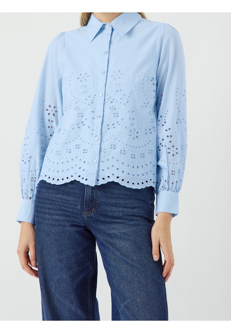 Femme portant un chemisier bleu clair boutonné avec broderie florale ajourée et ourlet festonné, associé à un jean bleu foncé.