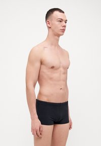 Čierne plavky s priliehavým strihom, hladkou textúrou a elastickým pásom, predvedené na stojacom mužskom modeli.