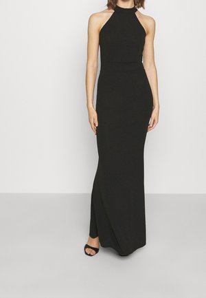 Kobieta ubrana w czarną, bezrękawnikową suknię na szyję, sięgającą do ziemi, z bocznym rozcięciem, oraz czarne sandały na obcasie, stojąca na tle jednolitego tła.