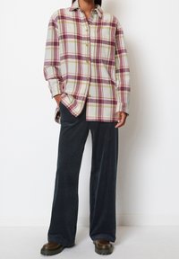 Plaid shirt in beige, burgundy, and green; features a button front and long sleeves. Paired with dark corduroy pants and brown footwear.