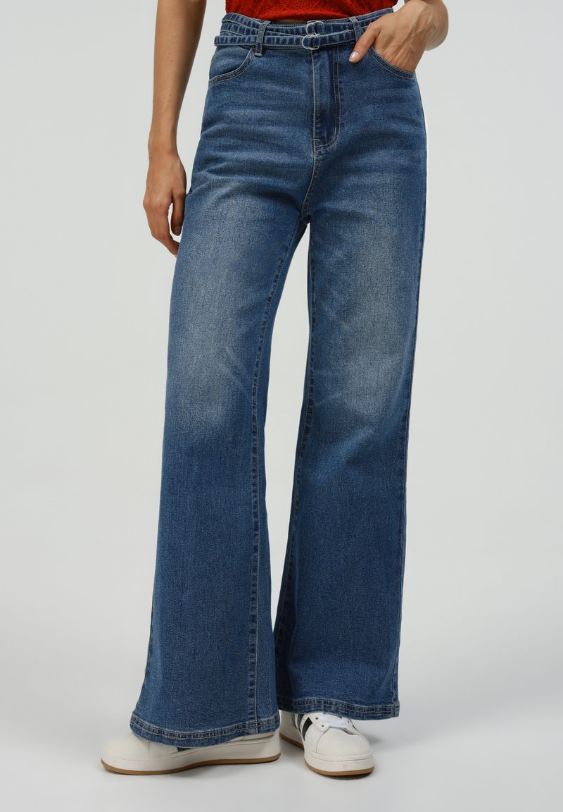 Wide-leg blue denim jeans with a high waist, front pockets, and a faded texture; paired with white sneakers for a casual look.