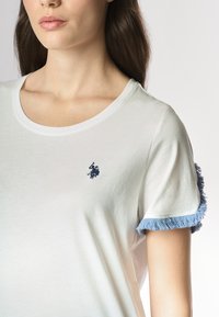 T-shirt bianca in cotone con scollo rotondo, maniche corte con dettagli in frange azzurre e logo ricamato blu sul petto.