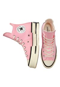 Baskets montantes en toile rose avec semelles en caoutchouc noires, lacets blancs et un écusson logo circulaire à l'extérieur. Présentent des motifs texturés.