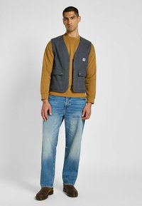 Gilet in tessuto grigio con cerniera frontale, due ampie tasche davanti, indossato sopra un felpa marrone, abbinato a jeans azzurri chiari e stivali scuri.