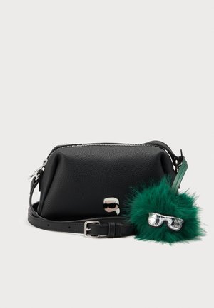Sac bandoulière en cuir noir texturé avec boucle argentée, fermeture éclair, petit détail métallique en forme de visage et porte-clés vert duveteux attaché avec lunettes de soleil miroir.