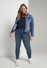 Chaqueta de mezclilla, azul claro, con bolsillos y botones, combinada con una blusa de azul oscuro. Jeans ajustados de mezclilla azul y zapatillas blancas completan el look.