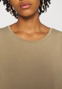 T-shirt kaki à encolure arrondie et texture douce. Il présente des coutures visibles et est associé à de grandes boucles d'oreilles en or.