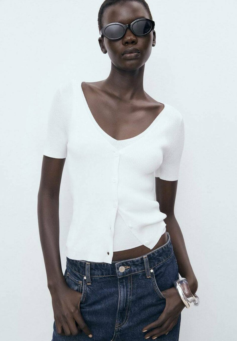 White ribbed short-sleeve cardigan with a v-neck and side buttons, paired with dark blue denim pants and clear bangle. Black sunglasses.