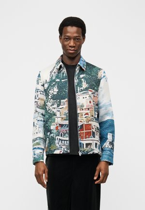 Hombre con una chaqueta con cremallera y un estampado colorido de un paisaje urbano, de pie contra un fondo blanco liso, mirando hacia adelante con expresión neutral.