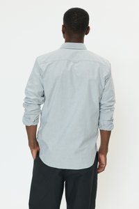 Camisa de manga larga gris claro con cuello abotonado y mangas remangadas. Presenta una tapeta en la parte trasera y un dobladillo redondeado.