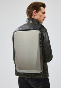 Sac à dos rigide gris avec une surface lisse, de forme rectangulaire, zips latéraux, et un panneau de contrôle comportant des ports et des boutons.
