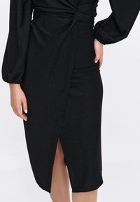 Schwarzes Midi-Kleid aus schimmerndem Stoff, mit einer geknoteten Vorderseite, langen Ärmeln und einem seitlichen Schlitz für zusätzlichen Flair.