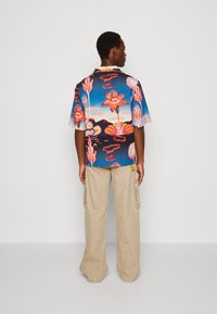 Cămașă cu mânecă scurtă de culoare albastră strălucitoare, cu modele florale în nuanțe de roz și portocaliu, asortată cu pantaloni cargo ușori bej deschis, dotati cu buzunare laterale.