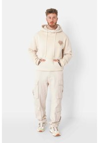 Beiger Kapuzenpullover mit Kordeln und Herzdesign, kombiniert mit passenden Cargo-Hosen mit seitlichen Taschen. Die Schuhe sind hellfarbene Laufschuhe.