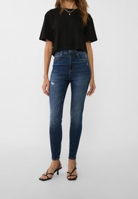 Jeans de tiro alto en azul oscuro con un ajuste ceñido y un ligero desgastado, combinados con una camiseta negra cropped y tacones negros con tiras.