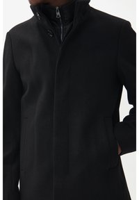 Svart ullblandad kappa med hög krage, dragkedja framtill och två sidfickor. Slät textur med minimalistisk design och inga mönster.