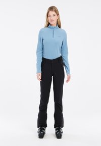 Maglione in pile azzurro chiaro con chiusura a zip abbinato a pantaloni da sci neri. Stivali da sci neri ai piedi. Texture liscia, design aderente, marchio discreto sulla parte superiore.