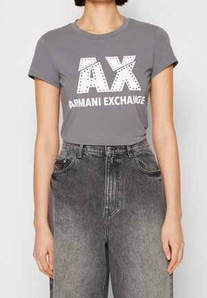 Woman wearing fitted grey Armani Exchange t-shirt with studded "AX" logo and high-waisted faded black jeans, standing against plain background.