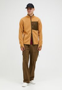 Beige Fleecejacke mit einer dunkelgrünen Brusttasche, kontrastierenden Reißverschlüssen, kombiniert mit braunen Cargo-Hosen und weißen Schuhen. Schwarze Mütze auf dem Kopf.