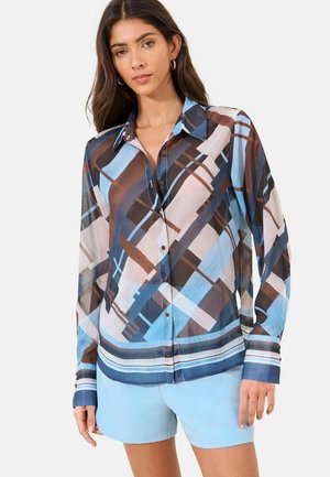 Frau trägt ein langärmliges Hemd mit Knöpfen in abstrakten geometrischen Mustern in Blau, Braun und Weiß sowie hellblaue Shorts vor weißem Hintergrund.