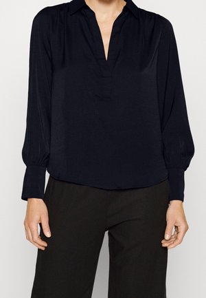 Kvinde iført en sort V-halsbluse med lange ærmer og løse manchetter, kombineret med sorte bukser, vist fra nakke til hofter.