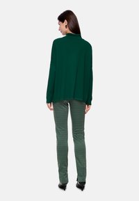 Grünes Langarmoberteil mit lockerer Passform, kombiniert mit gemusterten grünen Hosen mit geometrischem Design und schwarzen Absatzschuhen.