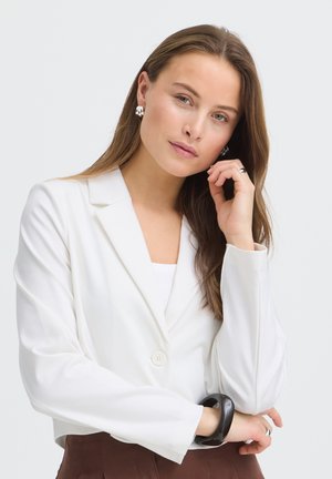 Jonge vrouw met lang bruin haar, een witte blazer, zilveren oorbellen en een zwarte armband dragend, met haar hand steunend op haar kin tegen een witte achtergrond.