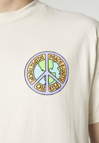 Béžové bavlněné tričko s kulatým grafickým designem znázorňujícím symbol míru a ilustraci Země. Text zní "SVĚTOVÝ MÍR A JEDNOTA, POSLUŠEJ."