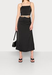 Completo nero in due pezzi composto da un top corto in satin con spalline sottili e una gonna maxi a vita alta, abbinato a sandali flat neri e una borsa piccola.